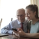 Young positive lady showing photos on smartphone to senior man while sitting at laptop